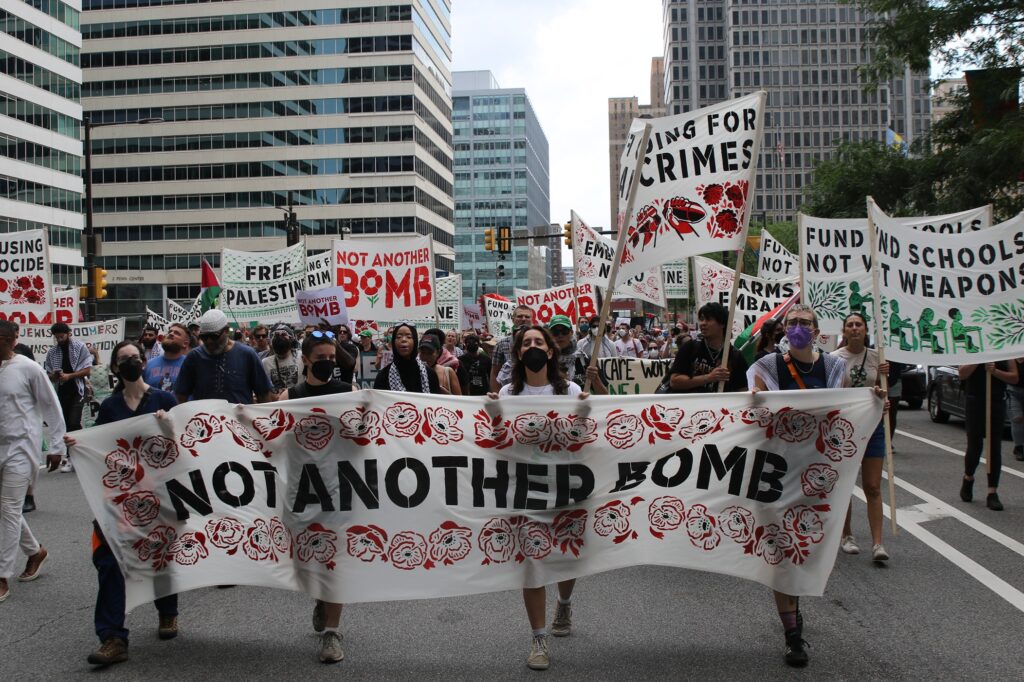 protesters hold signs in the street. The biggest reads not another bomb