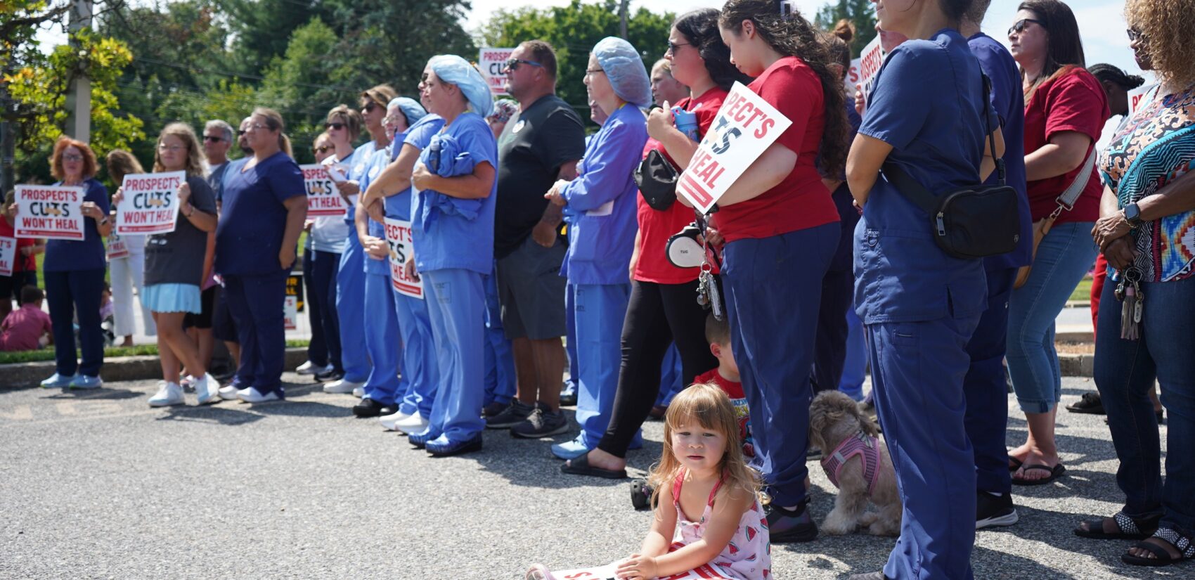 Dozens of health care professionals stand holding signs, while a child sits