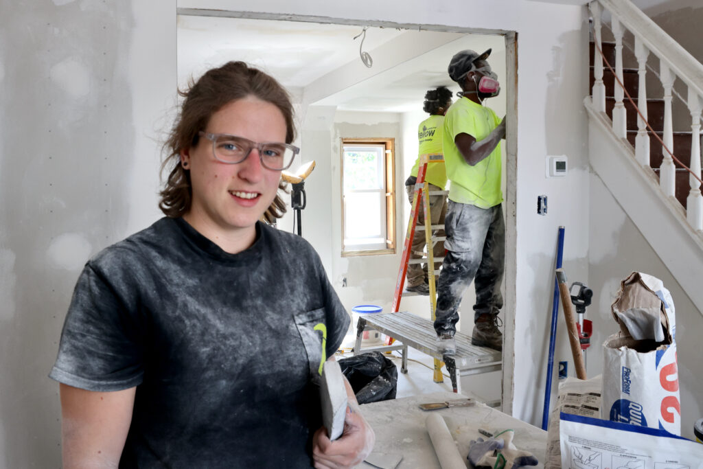 Zee Whitmer smiles to the camera while workers work on a house behind her