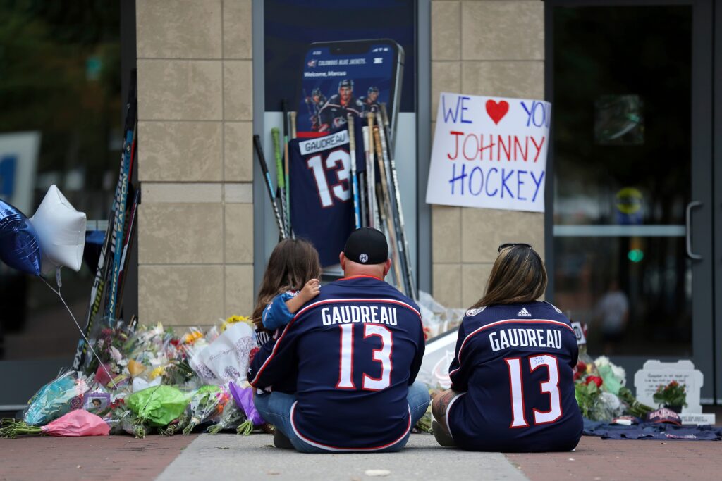 Johnny-Gaudreau-memorial-ohio Two people wearing Gaudreau jerseys sit and hold a kid at the memorial