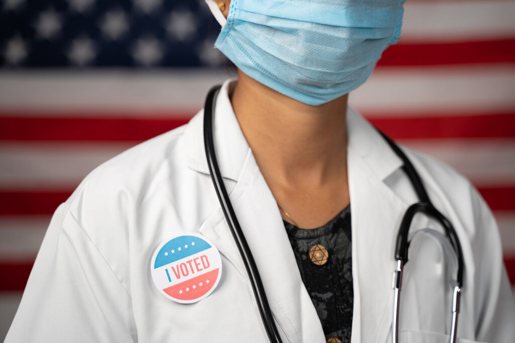 Doctor Or Nurse Medical Mask And Stethoscope Wearing Placing I V Doctor or nurse medical mask and stethoscope wearing placing I voted sticker to her apron - concept of US election with US flag as background