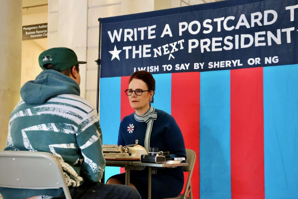 2024 11 05-e lee-andrew thomas-sheryl oring-philadelphia central library-postcards to the president Artist Sheryl Oring listens as Andrew Thomas tells her what he wants to say to the next president of the United States. Oring set up shop at Philadelphia's Central Library and typed the postcard on a vintage typewriter.