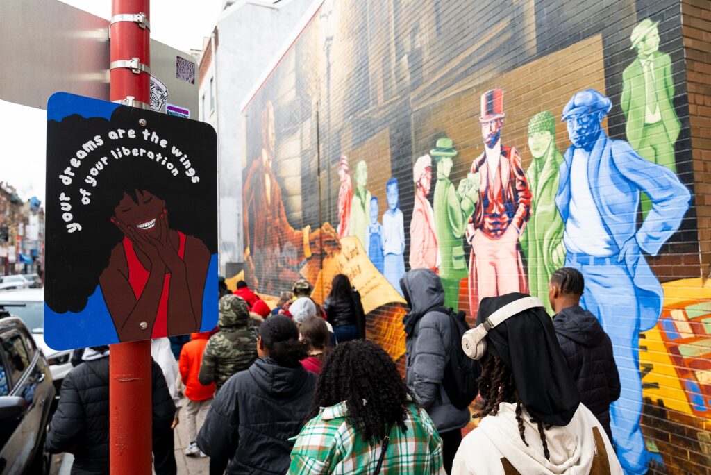 afromation-avenue-2024-philadelphia-2 people walk by a mural and a sign reading your dreams are the wings of your liberation