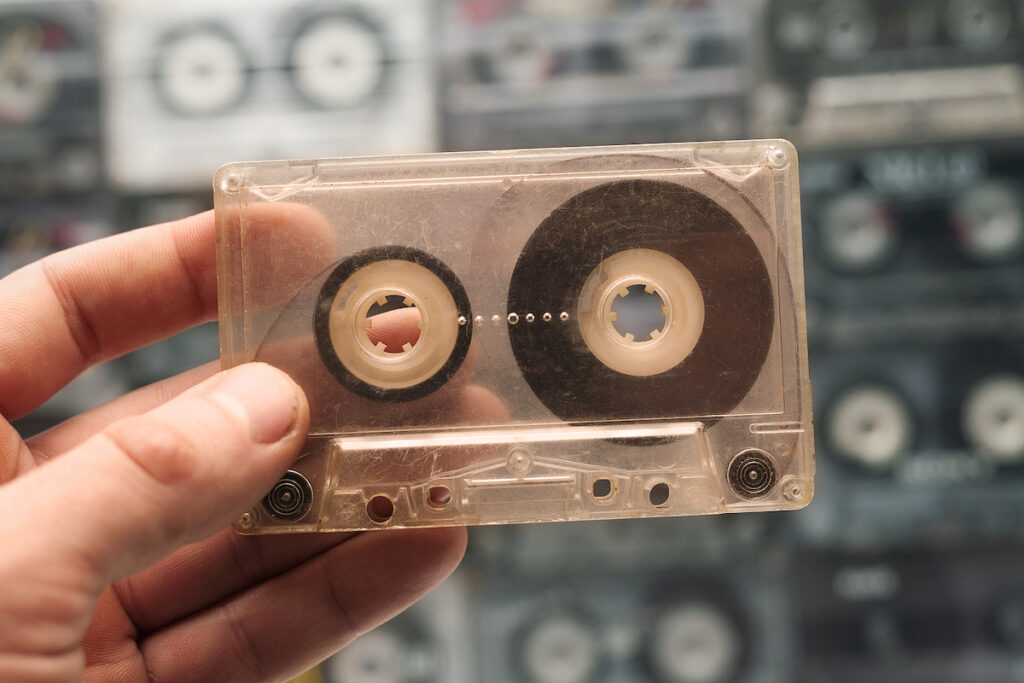 Hand Holds The Cassette. Retro Tape Cassette Music. Old Music. A person holds one old cassette tape out of many. (Bigstock/Tik.Tak)