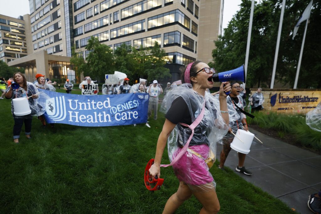 Health Care Advocates Risk Arrest Protesting Care Denials At UnitedHealthcare A woman speaking into a megaphone during a protest march, with people carrying a large banner behind her
