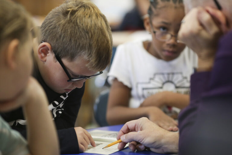 Student reading from a book with the help of a teacher