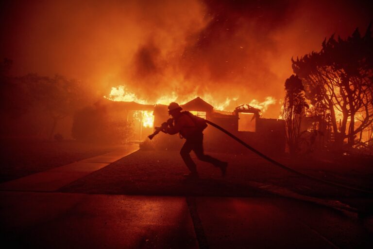 A firefighter dragging a fire hose in front of a house completely engulfed in flames