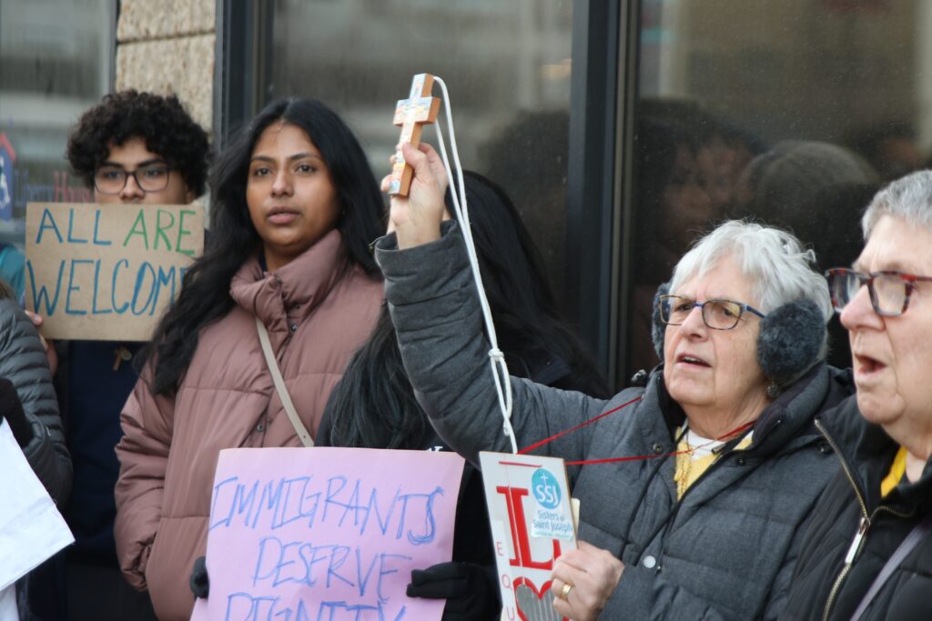 philadelphia-ice-raid-protest-2028-01-28-5 Protesters hold a cross and signs reading ALL ARE WELCOME and IMMIGRANTS DESERVE DIGNITY