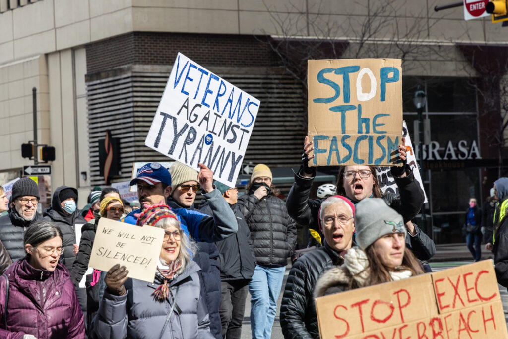 protesters walking, with a sign reading VETERANS AGAINST TYRANNY, another STOP THE FASCISM, and another reads WE WILL NOT BE SILENCED!