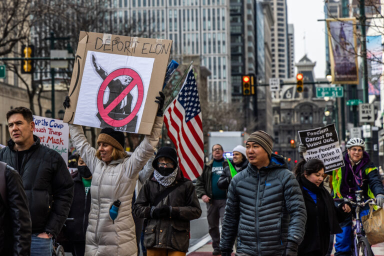 protesters walking in the street. a sign reads DEPORT ELON and shows his Nazi-like salute
