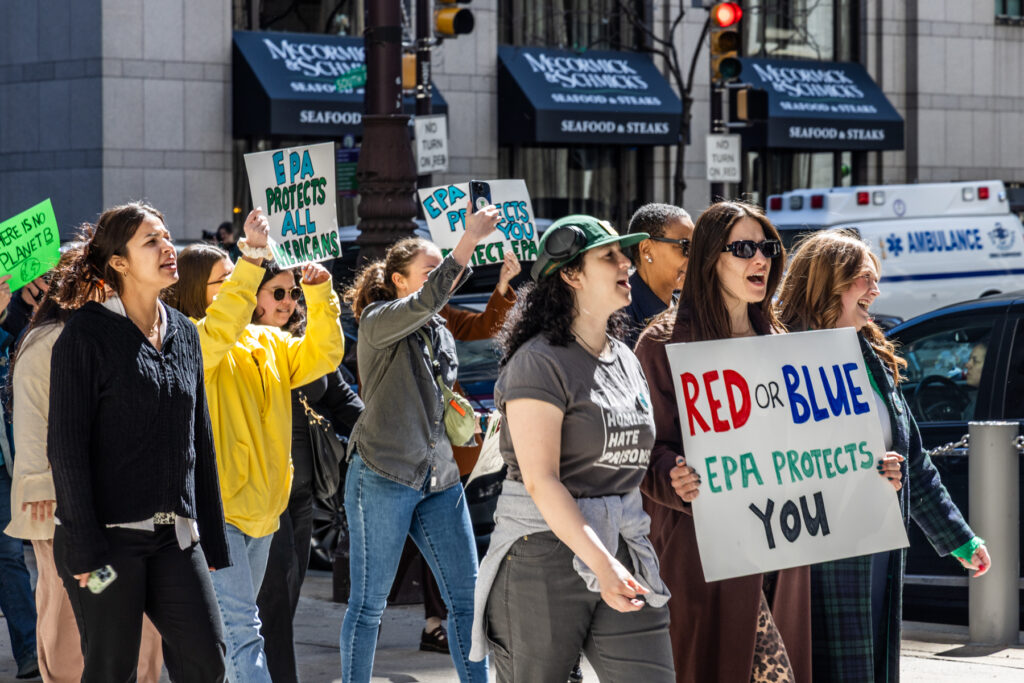 Protesters marching and carrying signs. One reads RED OR BLUE EPA PROTECTS YOU and another reads EPA PROTECTS ALL AMERICANS