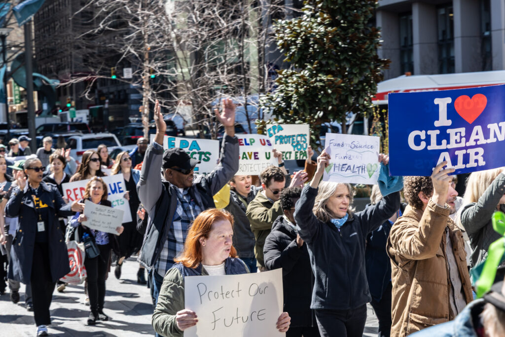 Protesters marching and carrying signs. One reads Protect Our Future and another reads Fund Science Fund Health