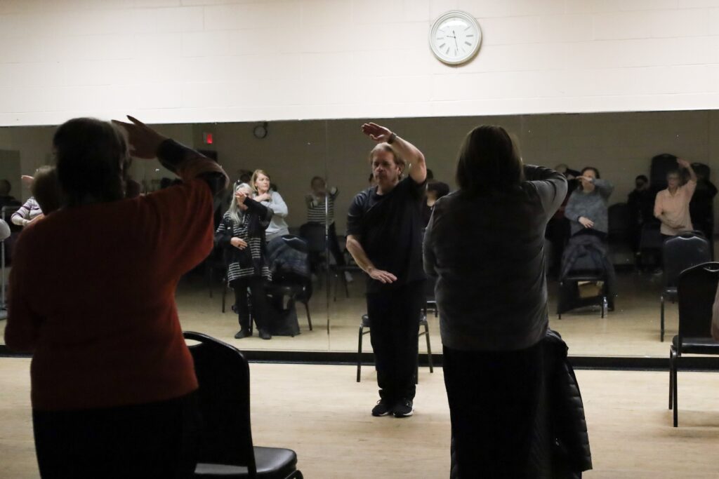 klein-life-n-leonard-2025-03-10-Retire 1 A man in the center of a large room does tai chi, and several people are doing it with him