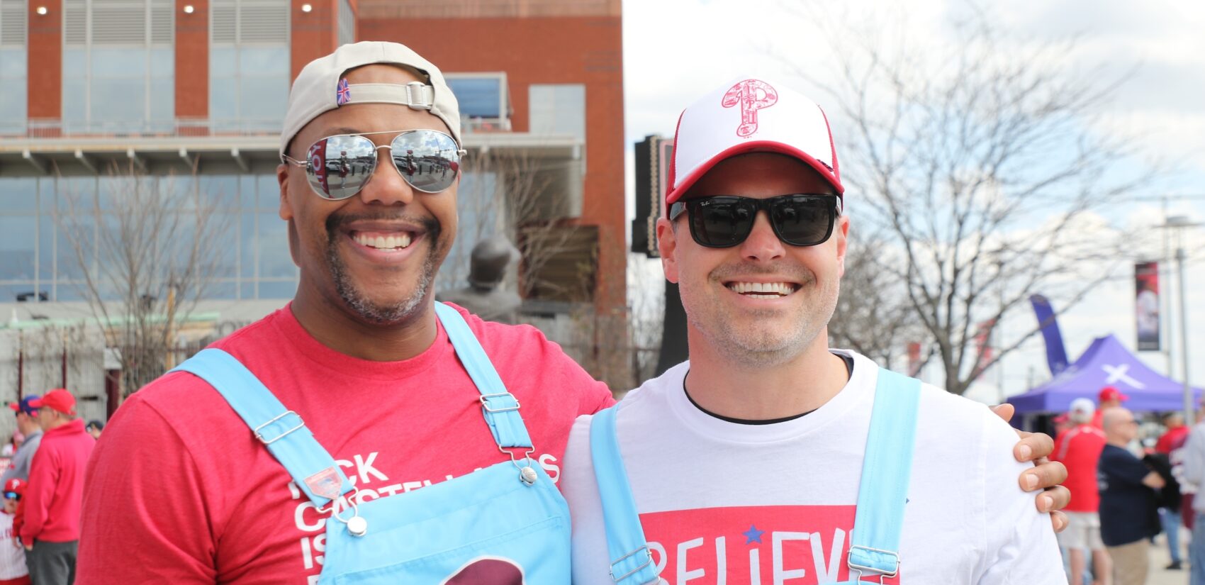 2 men wearing Phillies overalls and hats pose in front of the stadium