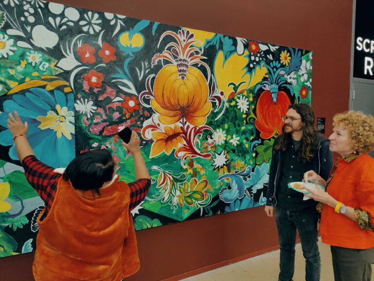 A woman gestures to a painting of flowers while a man and a woman look at it.