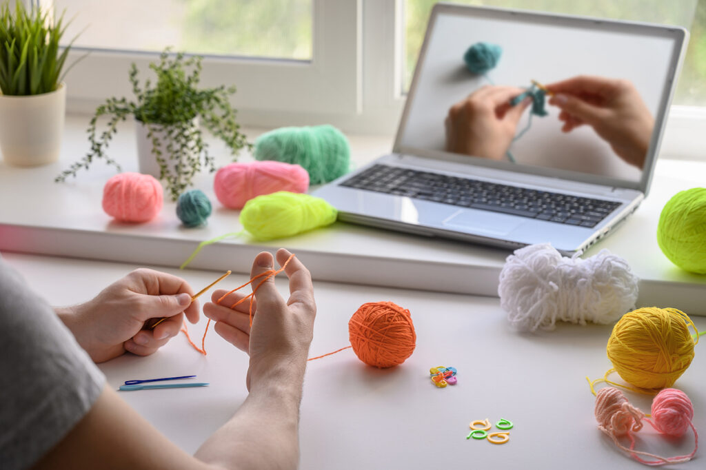 Selective Focus On The Hands Of A Man Sitting At The Table And Watching A Video Step By Step Guide O A man is crocheting and watching a tutorial video. (Ramil Gibadullin/Bigstock)