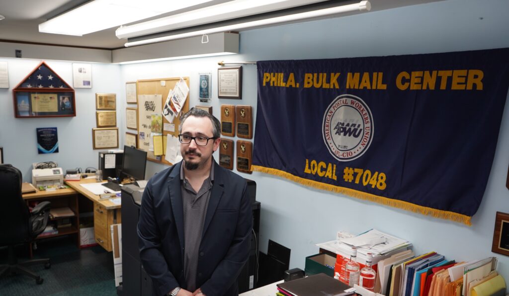 postal-workers-union-stand-against-trump-administration-KC-042525-02B.jpg James DeRidder standing in front of postal workers union flag