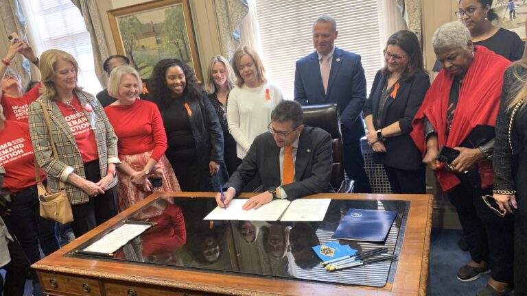 Matt Meyer signing a paper on his desk, surrounded by people
