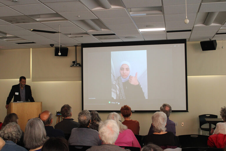 A woman from Northern Gaza is shown on a projector screen giving a thumbs up in front of a crowd