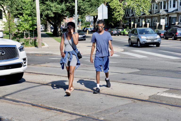 two people cross a crosswalk across Baltimore Avenue at Cobbs Creek Parkway