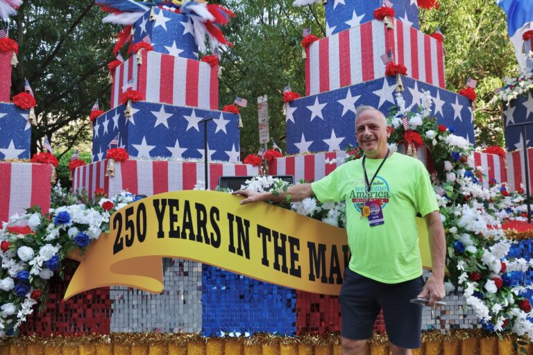 A man shows off a parade float designed to look like a large birthday cake with American flag colors.
