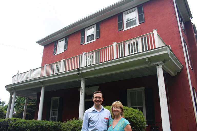 A man and a woman smile at the camera outside a red house
