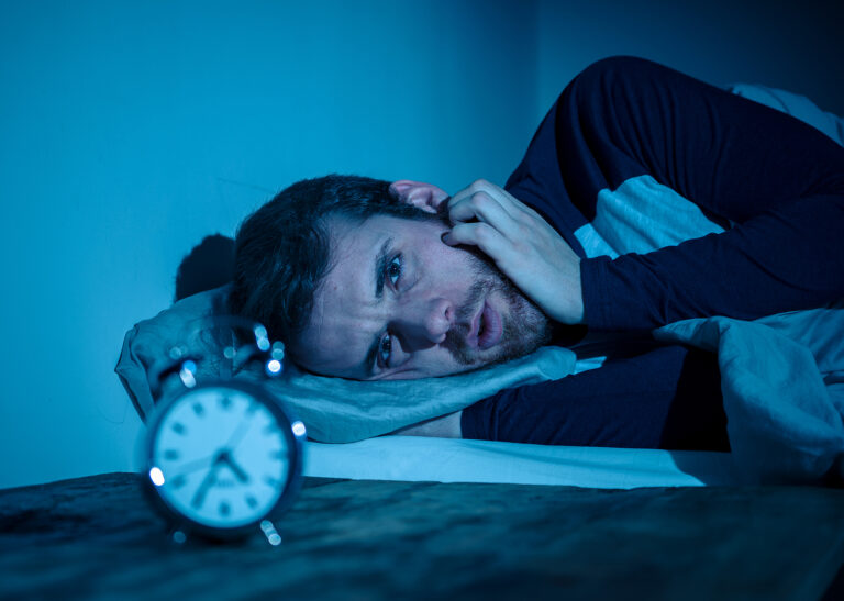 Sleepless and desperate young caucasian man awake at night not able to sleep, feeling frustrated and worried looking at clock suffering from insomnia in stress and sleeping disorder concept.