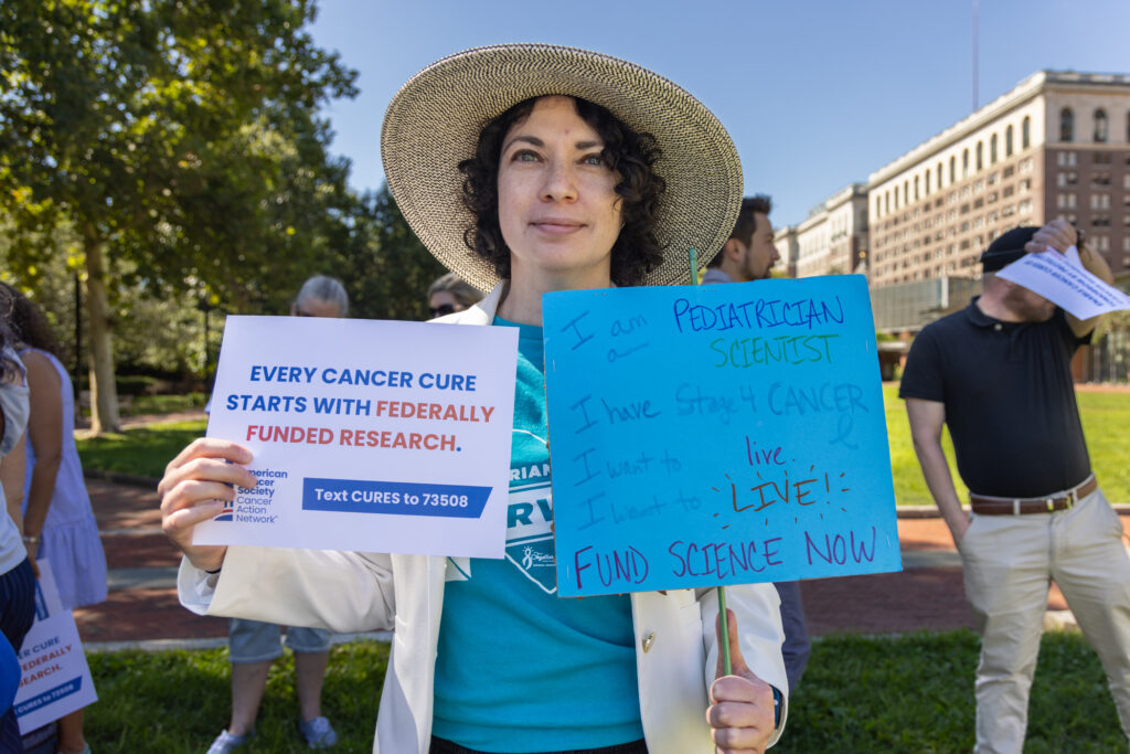 A pediatrician holds signs advocating for funding cancer research at Independence Hall