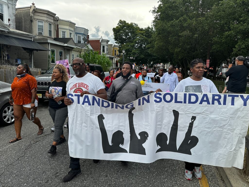2025-08-13-kburns-esiyah-dixon-waheed-remembered-2 Dozens marching through Camden holding a banner reading STANDING IN SOLIDARITY