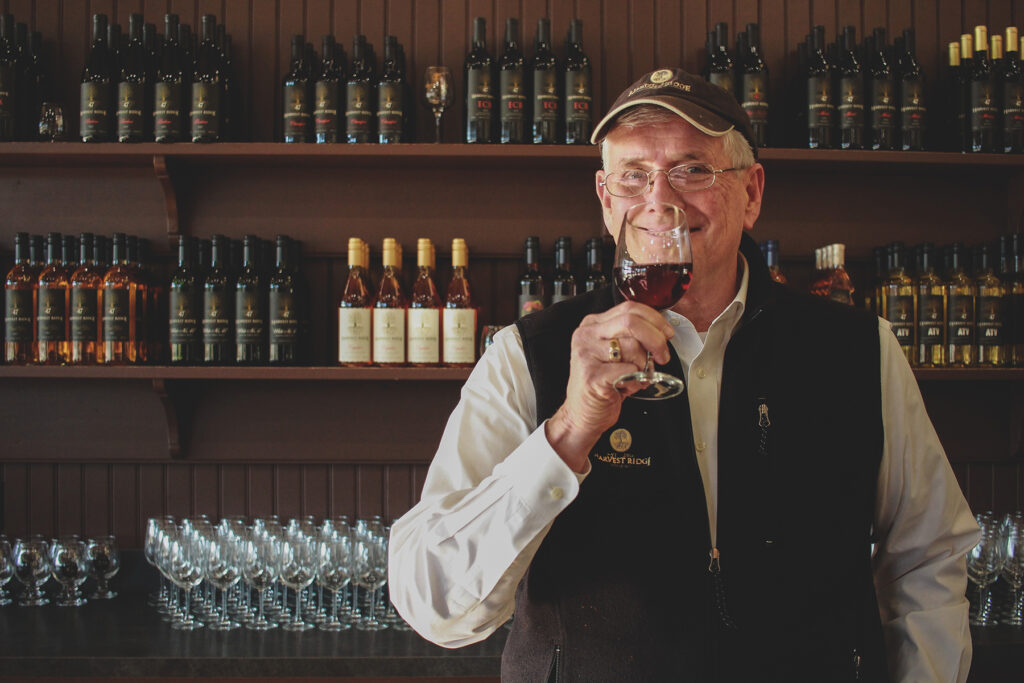 A man holds a glass of wine to his mouth while bottles of wine stand on shelves behind him