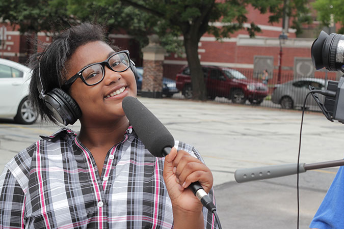 A young girl wearing headphones and holding a microphone outside smiling.