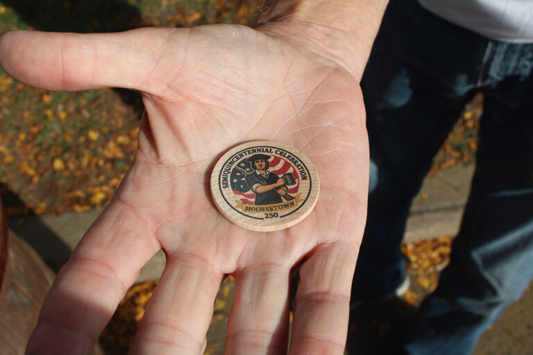 A hand holds a wooden coin celebrating Moorestown's Revolutionary War history.