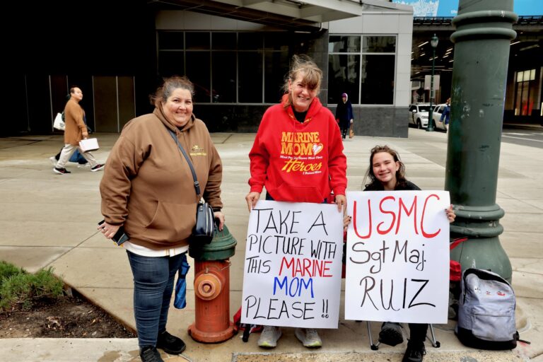 Three people with two signs, one reading TAKE A PICTURE WITH THIS MARINE MOM PLEASE!! and the other reading USMC SGT MAJ. RUIZ