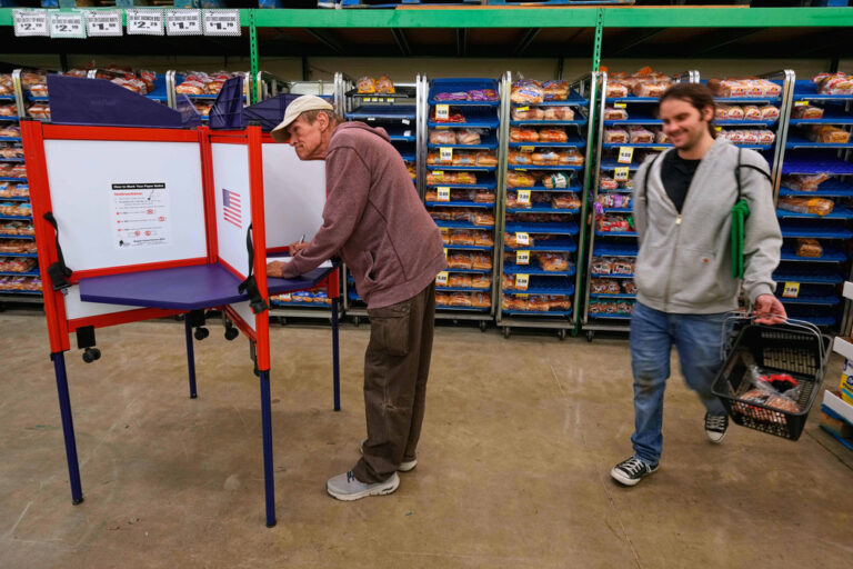 A person votes at a grocery store while another person walks by with a basket of food items