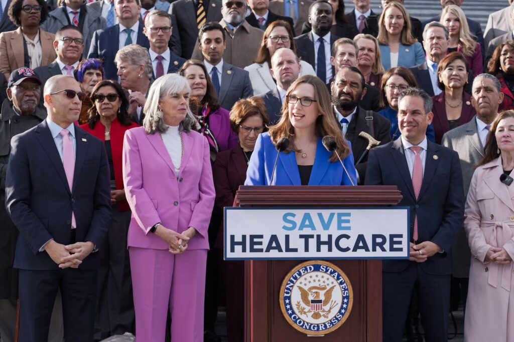 Government Shutdown Sarah McBride speaking at a podium surrounded by fellow Democrats