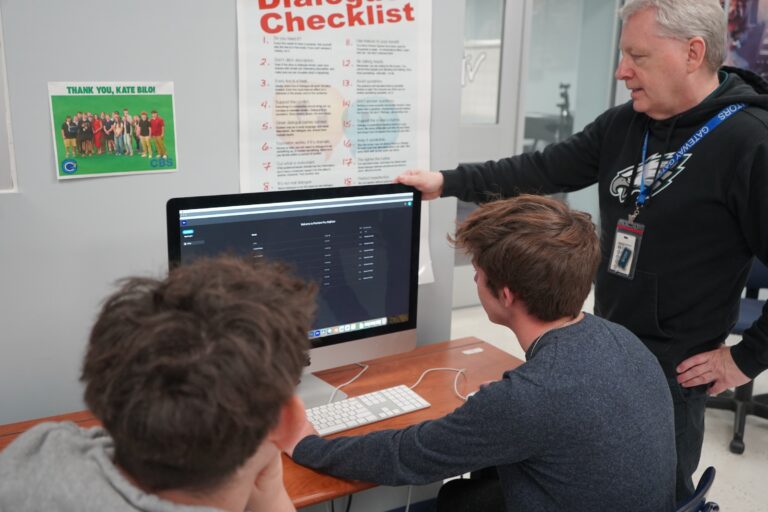A teacher looking intently at a computer monitor with two students