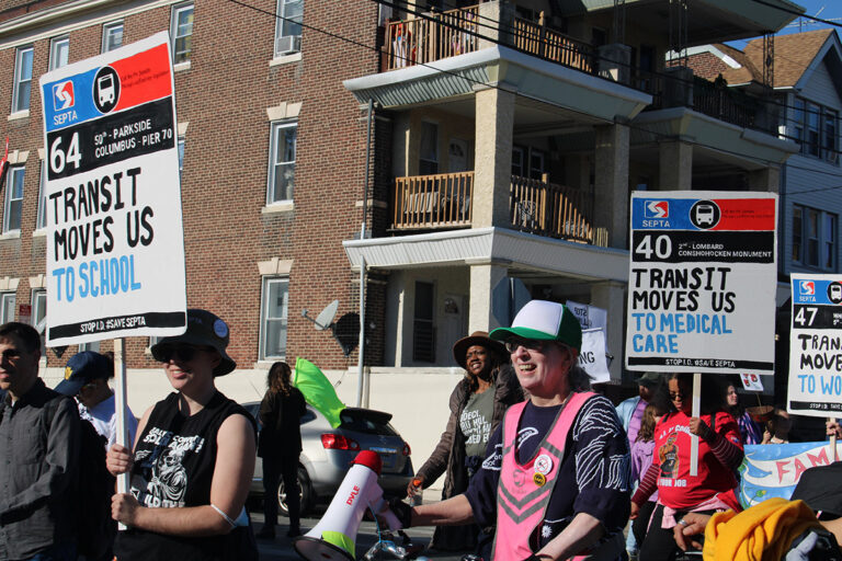 People hold up signs advocating for public transit at the 25th annual Peoplehood Parade in West Philadelphia
