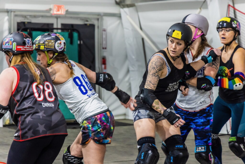 04 11 2025 k paynter roller derby stills-8.jpg Five women from the Brandywine Roller Derby team, wearing athletic clothes, kneepads, elbow pads, and wrist guards are in the middle of practicing for roller derby. They are also wearing helmets, which have a variety of colorful stickers on them.