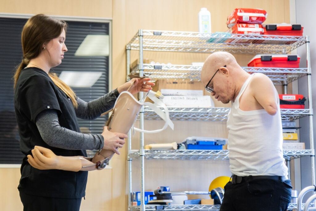 A doctor holds a prosthetic arm, and is tweaking something at the top, while Emmanuel looks on