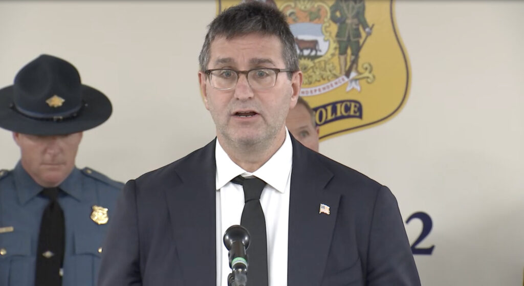 matt-meyer-dmv-wilmington-shooting Delaware Governor Matt Meyer addresses members of the media, speaking into a microphone at a podium, with a Delaware state trooper behind him.