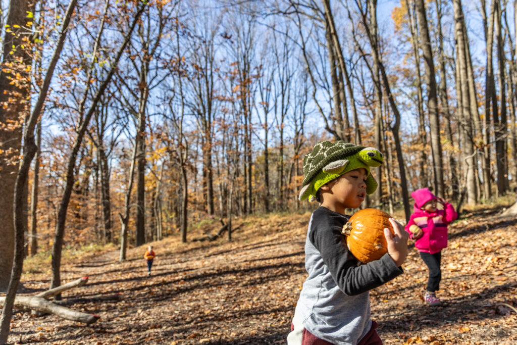 A child wearing a crochet turtle hat carries a pumpkin outside
