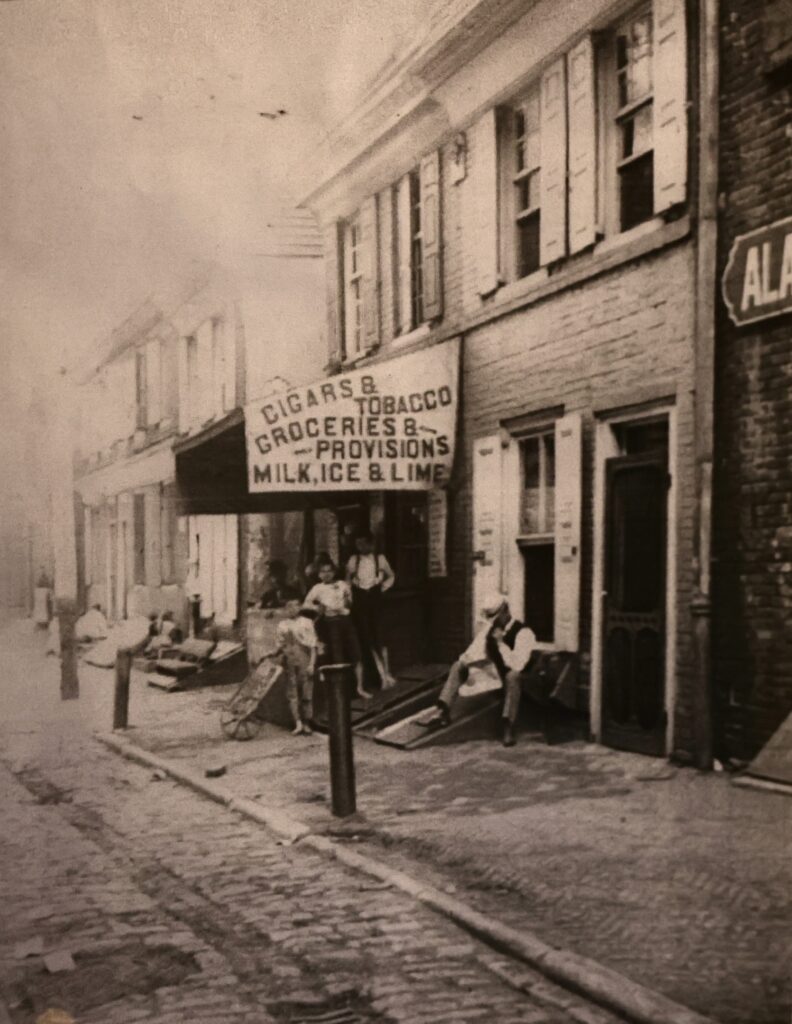 An archival black and white photo of a city block, with a sign for a store advertising cigars, groceries and tobacco