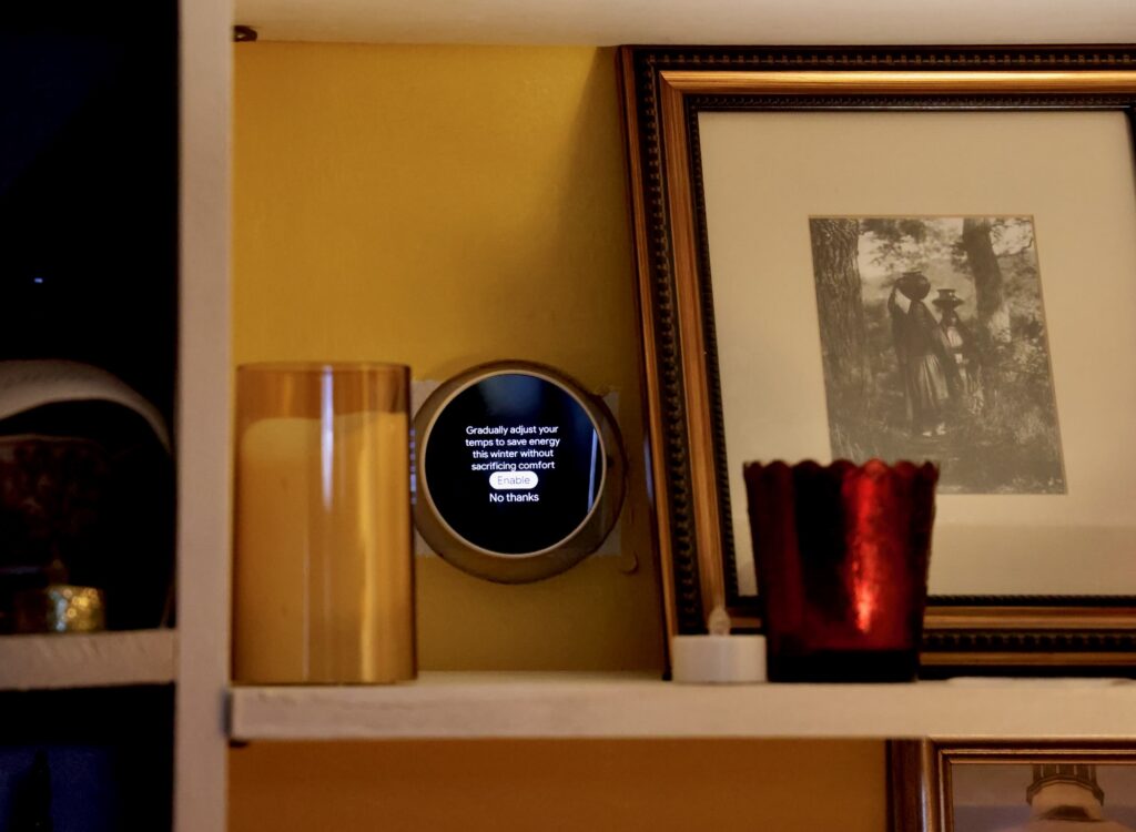 A smart thermostat is on the wall above a shelf with a candle and framed photo on it.
