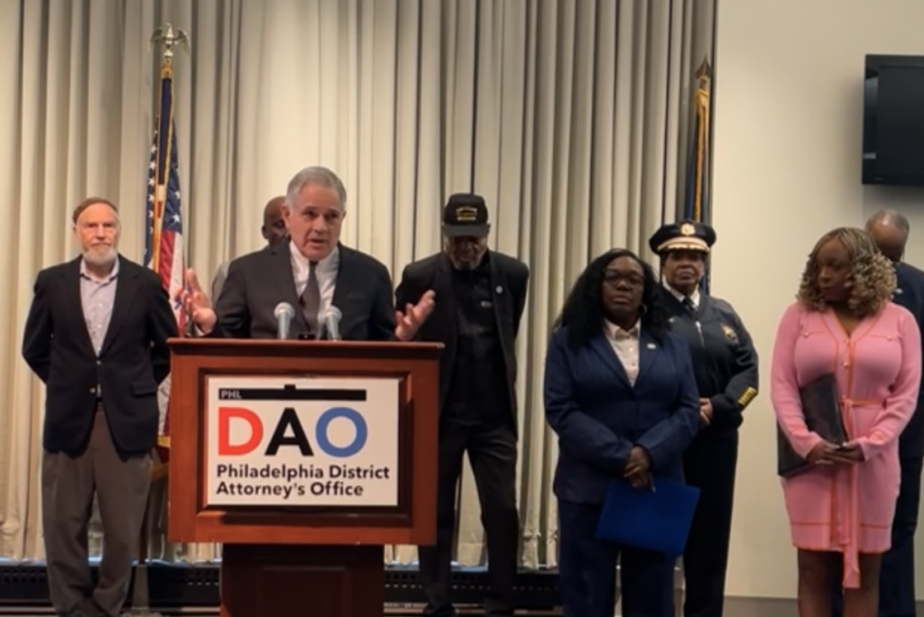 Larry Krasner speaking with many people behind him in front of a podium with a Philadelphia District Attorney's Office sign