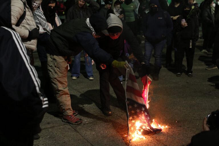 Two people burn an American flag in the middle of a crowd on the street