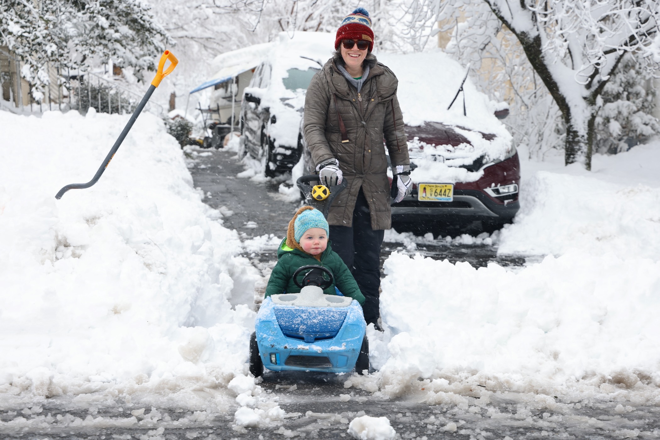 A toddler is in a blue child-sized car that is being pushed by its mother along a just-shoveled driveway after a massive snowstorm in Moorestown, New Jersey