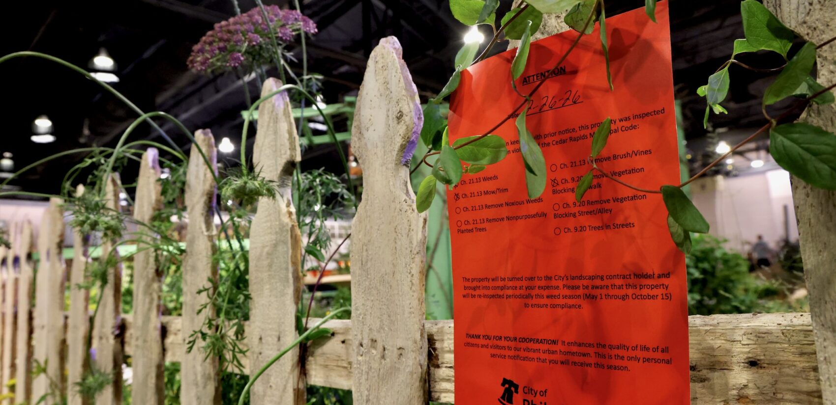 A notice ordering weeding, mowing and trimming is affixed to and overrun picket fence at the flower show