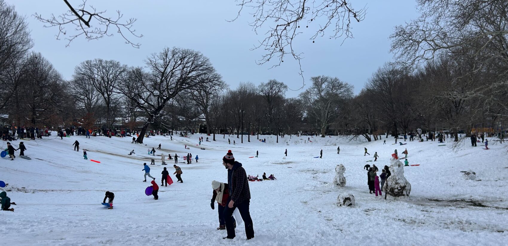 A snow filled park with many people sledding and building snowmen