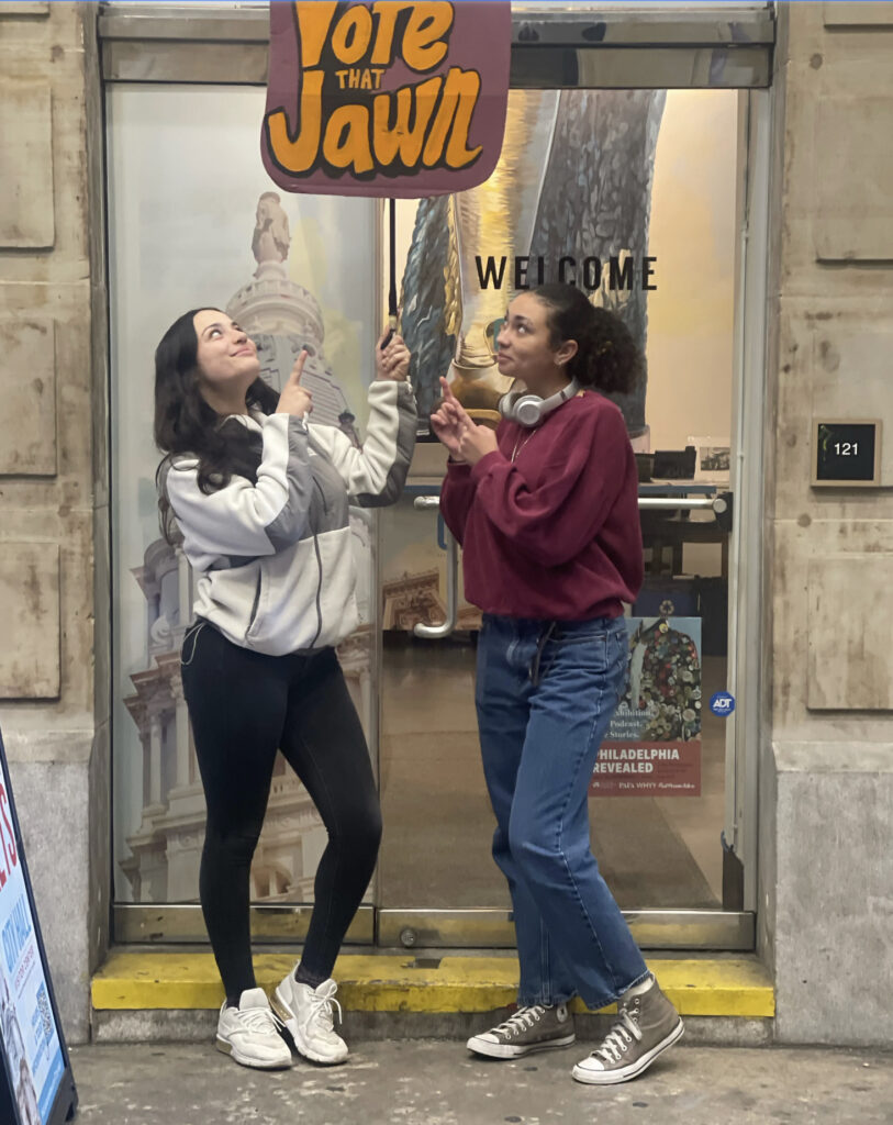 Two Vote That Jawn student members point to a sign above their heads that reads "Vote That Jawn"