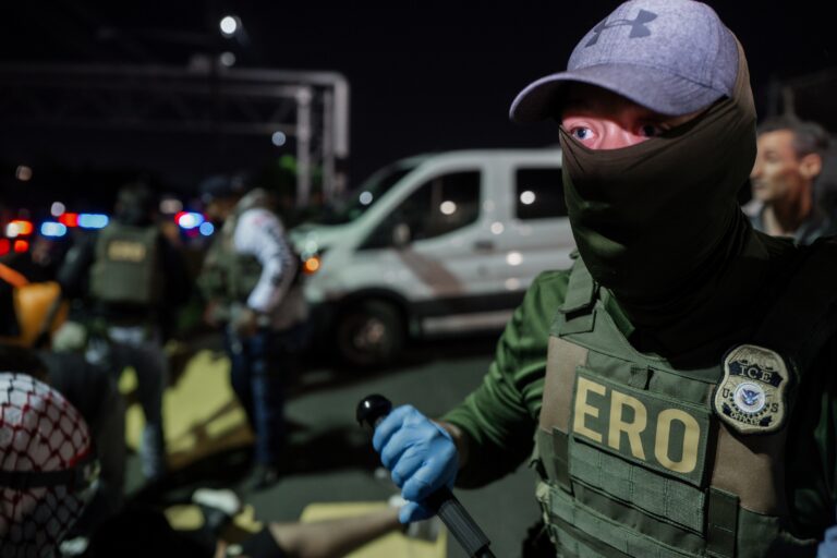A masked Immigration and Customs Enforcement agent holds a baton